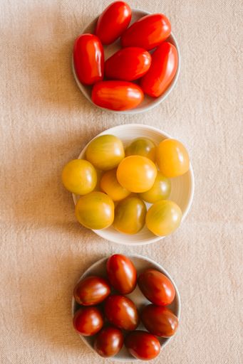 The flat lay of three bowls of yellow, red, and burgundy tomatoes on the light fabric background