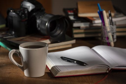 A closeup of a cup of coffee next to a notebook and pen, on a wooden study table