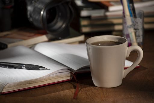 A closeup of a cup of coffee next to a notebook and pen, on a wooden study table