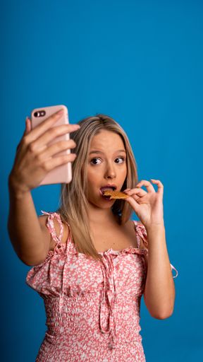 A vertical shot of a beautiful blond Caucasian woman eating a cookie while taking a selfie