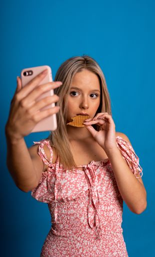 A vertical shot of a beautiful blond Caucasian woman eating a cookie while taking a selfie