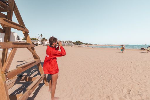 A male lifeguard with red jacket surveying the beach by looking through his binoculars