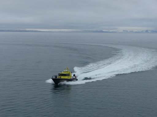 A drone view of a yellow boat sailing with foamy ripples behind it, entering the port of Spitsbergen, on a cloudy day