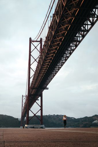 A vertical shot of a girl running by the river under the 25 de Abril Bridge in Lisbon, Portugal