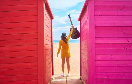 A backshot of a Caucasian teenager standing between colorful beach sheds and posing with an acoustic guitar - the concept of fun and leisure
