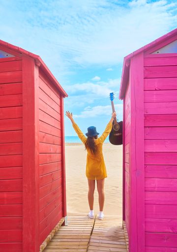 A backshot of a Caucasian teenager standing between colorful beach sheds and posing with an acoustic guitar - the concept of fun and leisure