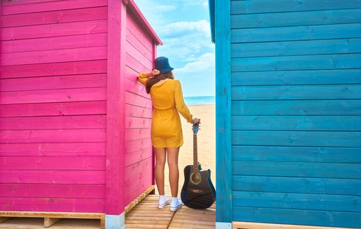 A backshot of a Caucasian teenager standing between colorful beach sheds and posing with an acoustic guitar - the concept of fun and leisure