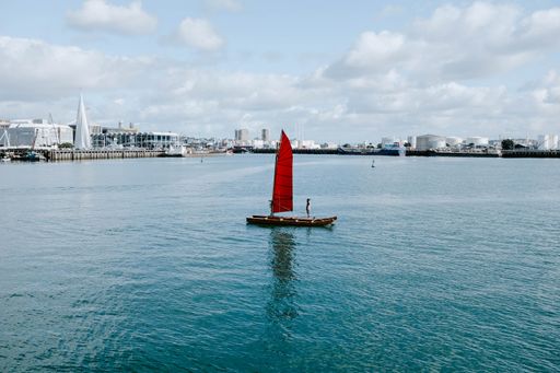 A small wooden red sailing boat floating on a sea with a port on the background under a cloudy sky