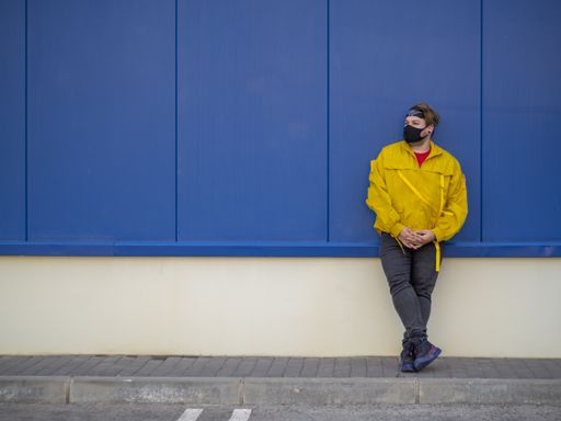 A male wearing a yellow jacket and a mask and posing by a blue wall