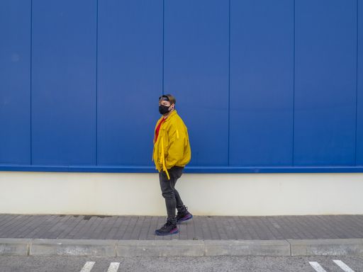 A male wearing a yellow jacket and mask posing by a blue wall