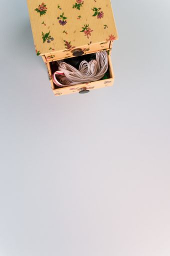 A vertical shot of a small wooden box with floral patterns and one drawer open, filled with threads, on a white surface