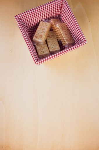 A vertical shot of crackers in plastic wrappers in a wooden basket with red checkered cloth all around