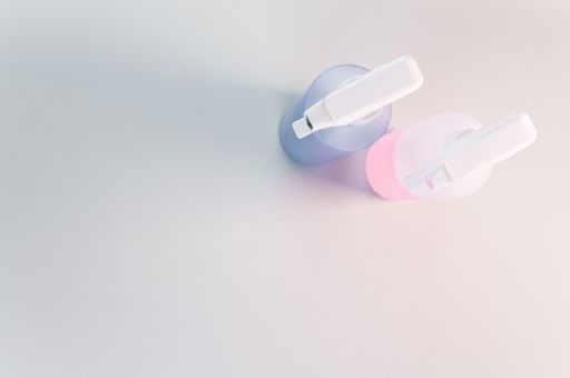 A high angle shot of two spray bottles with pink and purple liquid in them, on a white surface