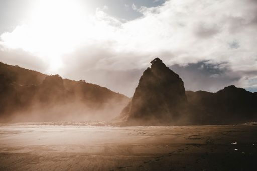 A landscape of a beach surrounded by rocky mountains under a cloudy sky during fog and sunset