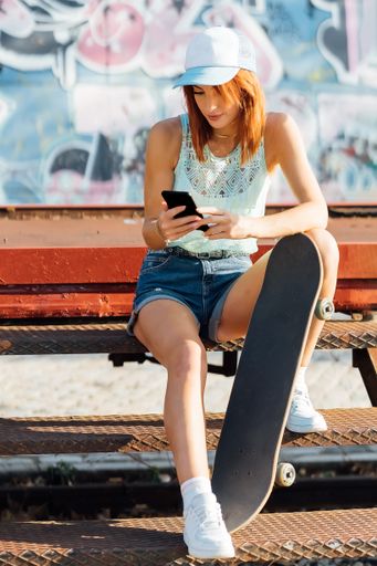 A vertical shot of a Caucasian young redheaded girl with skateboard looking at her mobile phone