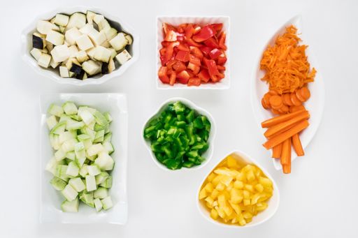 A top view of zucchini, carrots, peppers, and eggplants cut into cubes in white saucers on white table