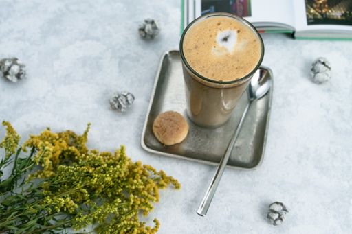 A top view of a coffee latte on a square saucer with a spoon, cookies, and crinkles on a white table
