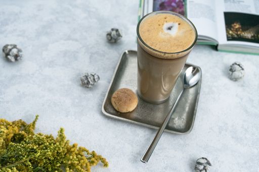 A top view of a coffee latte on a square saucer with a spoon, cookies, and crinkles on a white table