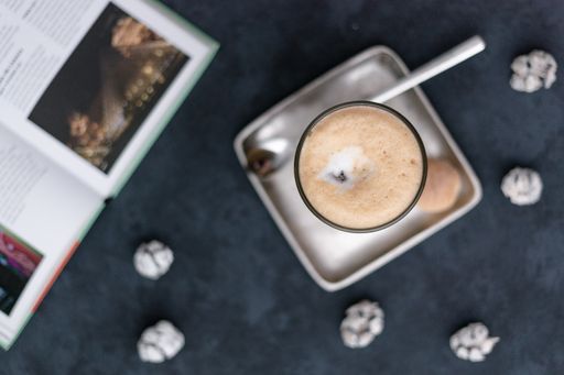 A top view of a coffee latte on a square saucer with a spoon, cookies, and crinkles on a black table