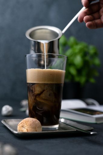 A vertical shot of hand adding milk to a coffee latte with cookies and spoon on a square saucer