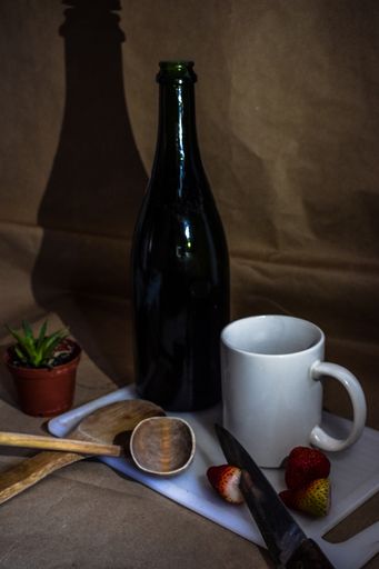 A vertical shot of fresh strawberries, knife, chopping board, mug, wooden spatula, bottle, and a plant on a surface