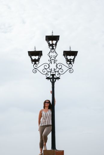 A vertical shot of a Caucasian woman leaning on a lamppost with a cloudy sky behind