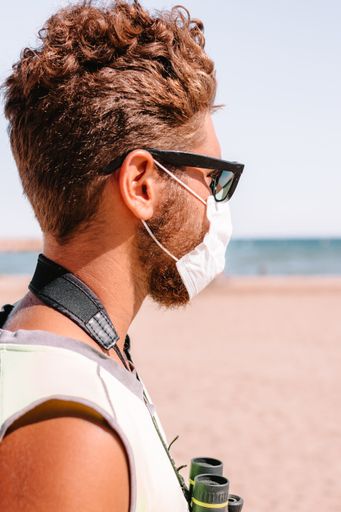 A vertical shot of a young caucasian male in a protective face mask with binoculars at the beach