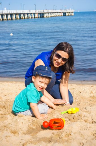 Woman with sunglasses playing with a toddler boy with plastic toys on sand at a beach close by the water.