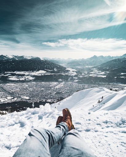 A vertical shot of a male's feet sitting on a snow covered cliff under the beautiful cloudy sky