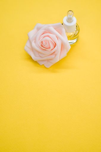 A vertical shot of essential oil next to a beautiful pink rose on a yellow surface
