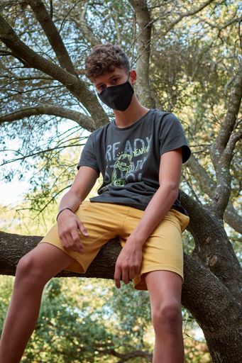 A young man sitting on a tree wearing a sanitary mask - concept of the new normal
