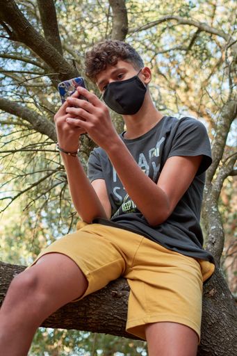 A young man sitting on a tree using his phone wearing a face mask-concept of the new normal