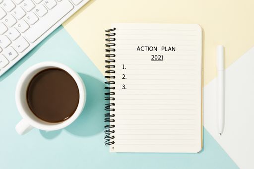 A flat lay shot of copybook with 'Action plan 2021' written on it, pen, cup of coffee and keyboard