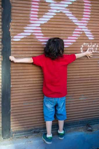 A vertical shot of a back view of a caucasian boy wearing a red shirt, hugging a shutter garage door