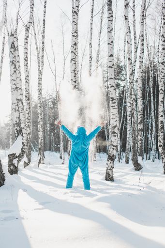 A vertical shot of a person in a fluffy blue costume standing with hands up in a forest in winter - back to childhood concept