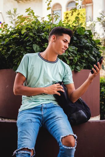 A vertical shot of a young Hispanic man sitting while texting through his phone