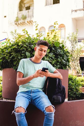 A vertical shot of a young Hispanic man sitting while texting through his phone
