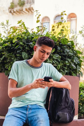 A vertical shot of a young Hispanic man sitting while texting through his phone