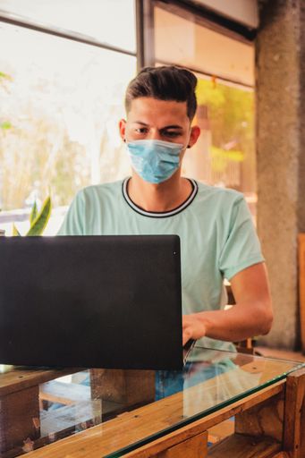 A vertical shot of a young Hispanic man with mask working on his laptop