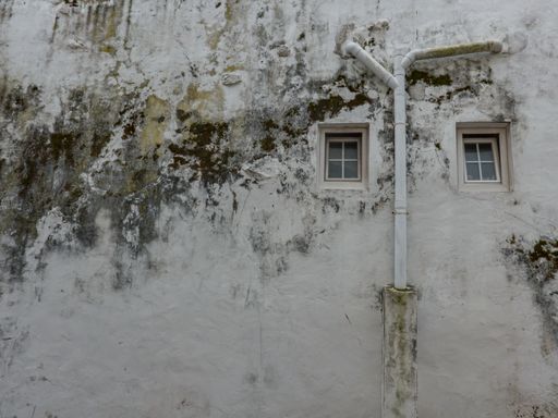 Grungy once white wall with dirt and mildew with two small windows and a drain pipe