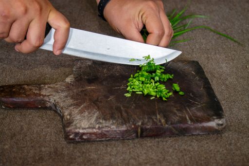 A closeup shot of a man's hand slicing onion chives on a wooden chopping board