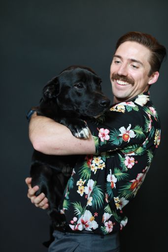 A vertical shot of a young Caucasian man in a floral shirt with a black Labrador retriever dog isolated on a black background