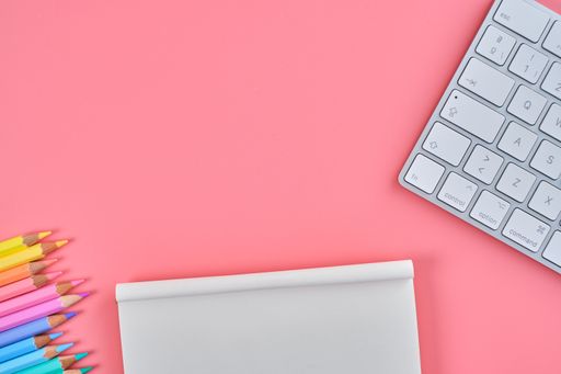 A top view of colored pencils, a notepad, and a keyboard on a pink background with copy space