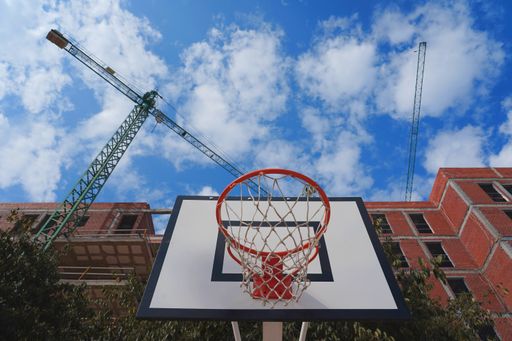 A low angle shot of construction site with a basketball ring, tower cranes, and buildings