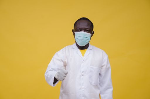 A closeup shot of a Nigerian man wearing a laboratory gown, gloves, and mask while doing thumbs up isolated on a yellow background