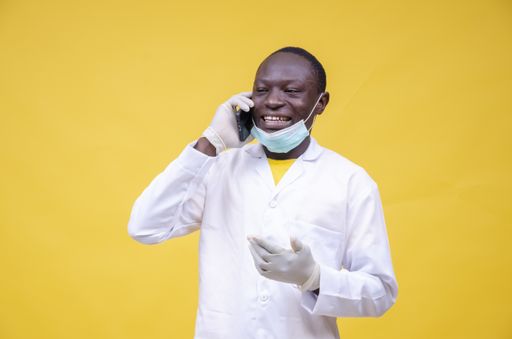 A closeup shot of a Nigerian man wearing a laboratory gown, gloves, and mask talking through a cellphone isolated on a yellow background