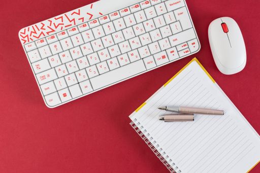 A top view of white keyboard, a pen, a notebook and s mouse on the red background