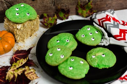 Green sugar cookie with eyeball decorations on a black plate. Fake skeleton hand is holding one. Cloth with pattern around plate and fall leaves.