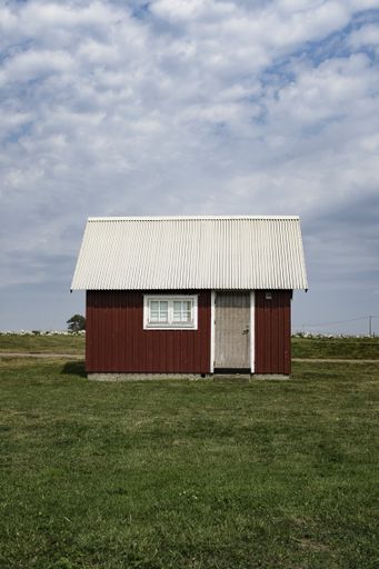 A wooden red barn with a white roof in a large green field with cloudy blue sky