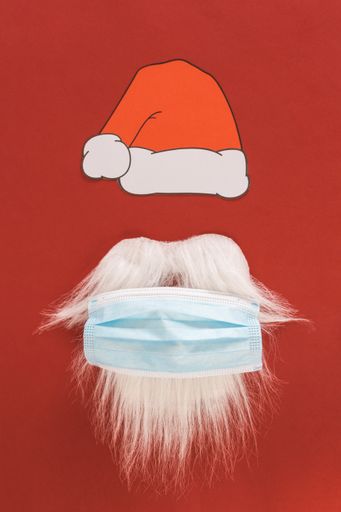 A closeup of a red Christmas hat, beard, and mustache of Santa Claus with a protective mask against the covid-19 pandemic on a red background